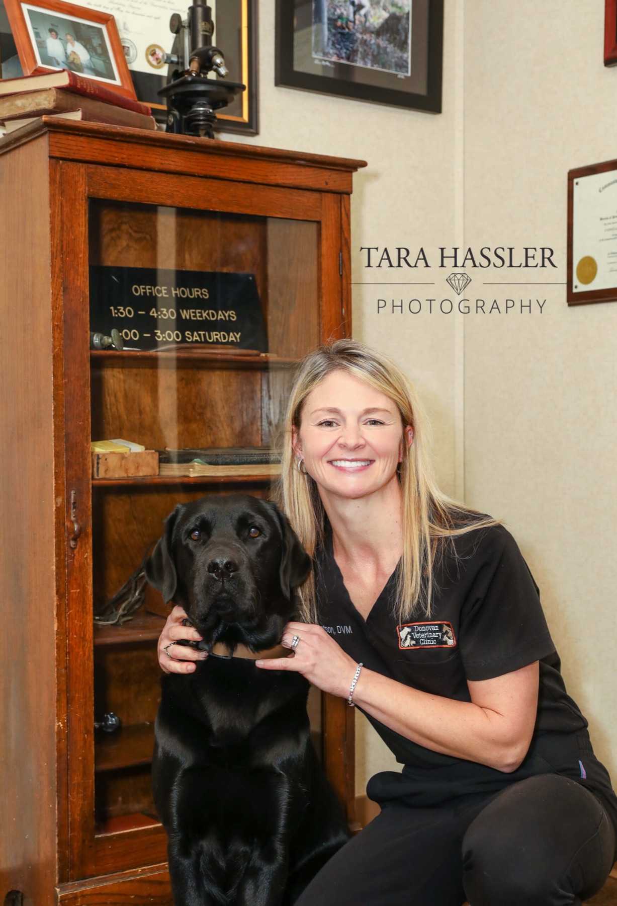 Dr. Connie Donovan Matson kneeling beside a black Labrador next to an antique wooden cabinet holding an old office-hours sign